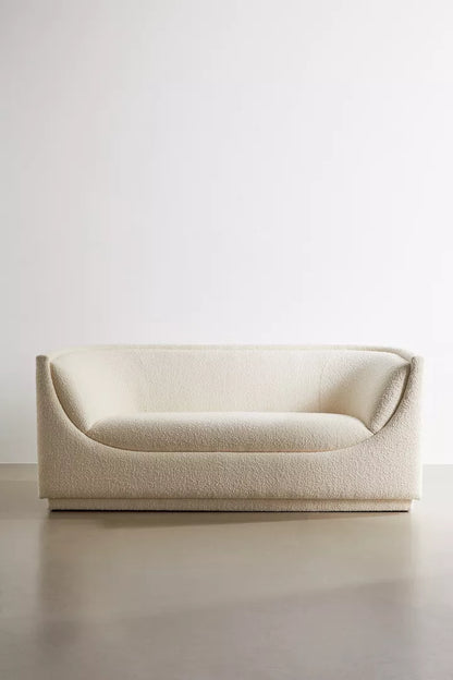 The Aria Sofa, with its minimalist cream tone and soft, curved design, is showcased against a plain white background on a smooth floor. It boasts textured fabric and exudes a modern aesthetic.