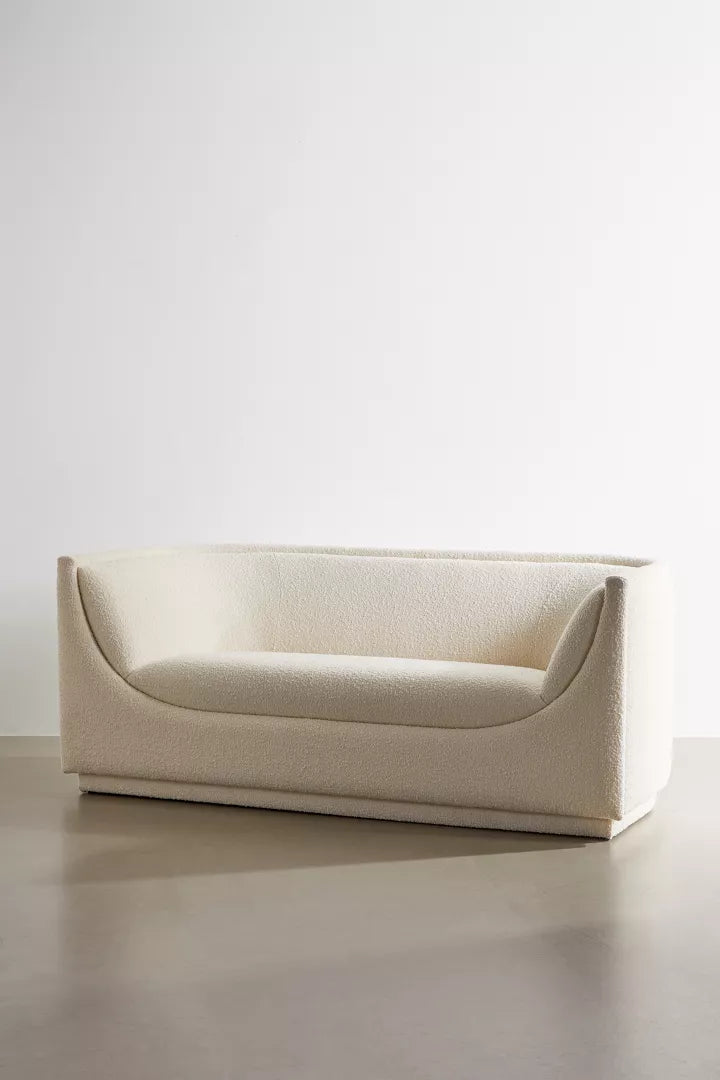 The Aria Sofa, showcasing a contemporary white design with elegant curved armrests and smooth textured fabric, is placed against a plain white wall on a light gray floor.