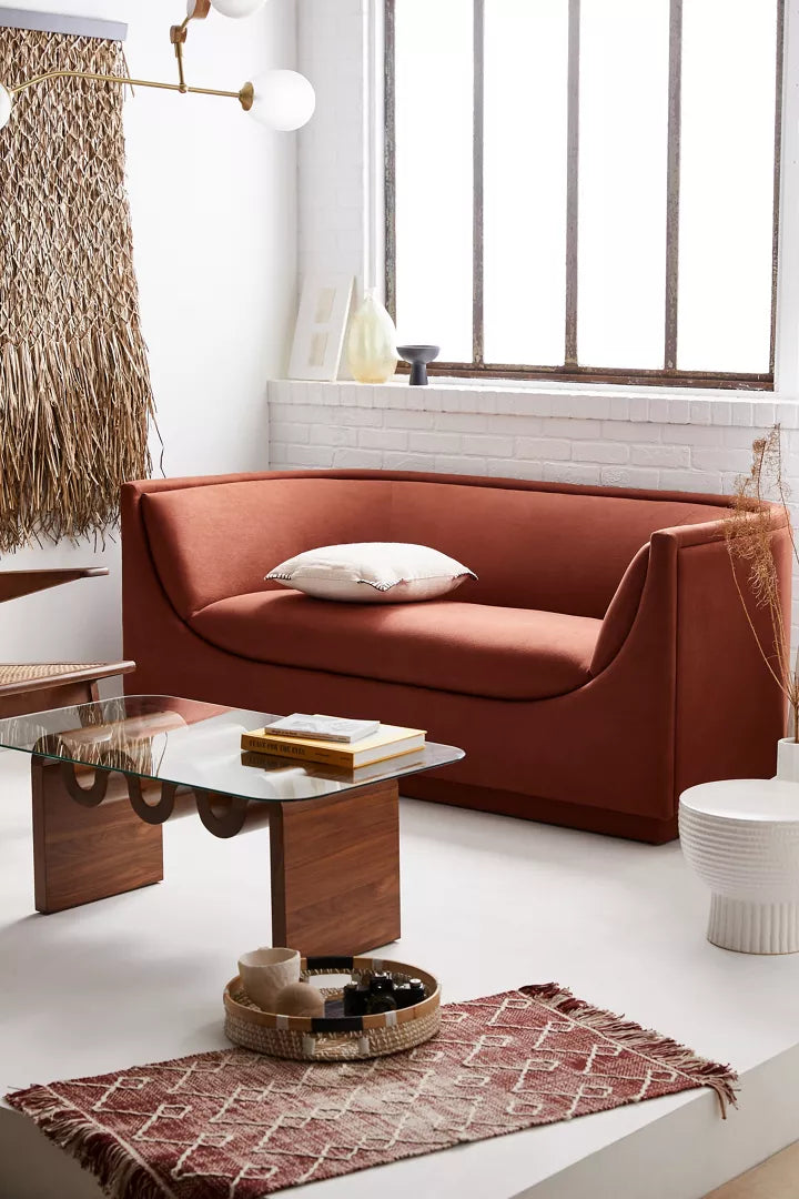 A modern living room features the Aria Sofa in a rust color, accompanied by a glass coffee table with wooden legs, a woven wall hanging, and a textured red rug. Sunlight streams through a large window, adding warmth to the minimalist decor. Decorative items are elegantly arranged on the table.