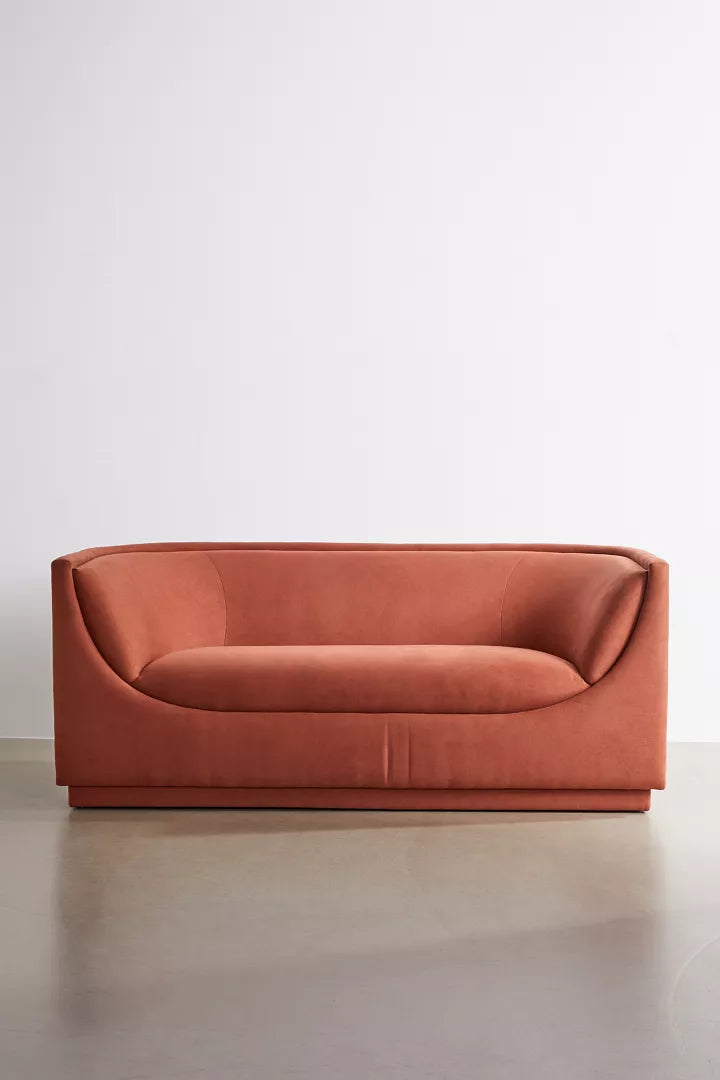 The Aria Sofa, a modern minimalist piece with smooth, rounded edges and a vibrant orange hue, is placed against a plain white wall. It boasts a sleek design with a seamless, cushioned back and rests elegantly on a polished concrete floor.