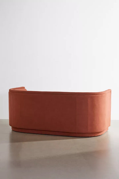 The Aria Sofa, with its modern curved design in orange, is placed on a minimalist floor against a plain white wall, highlighting its sleek and elegant style.