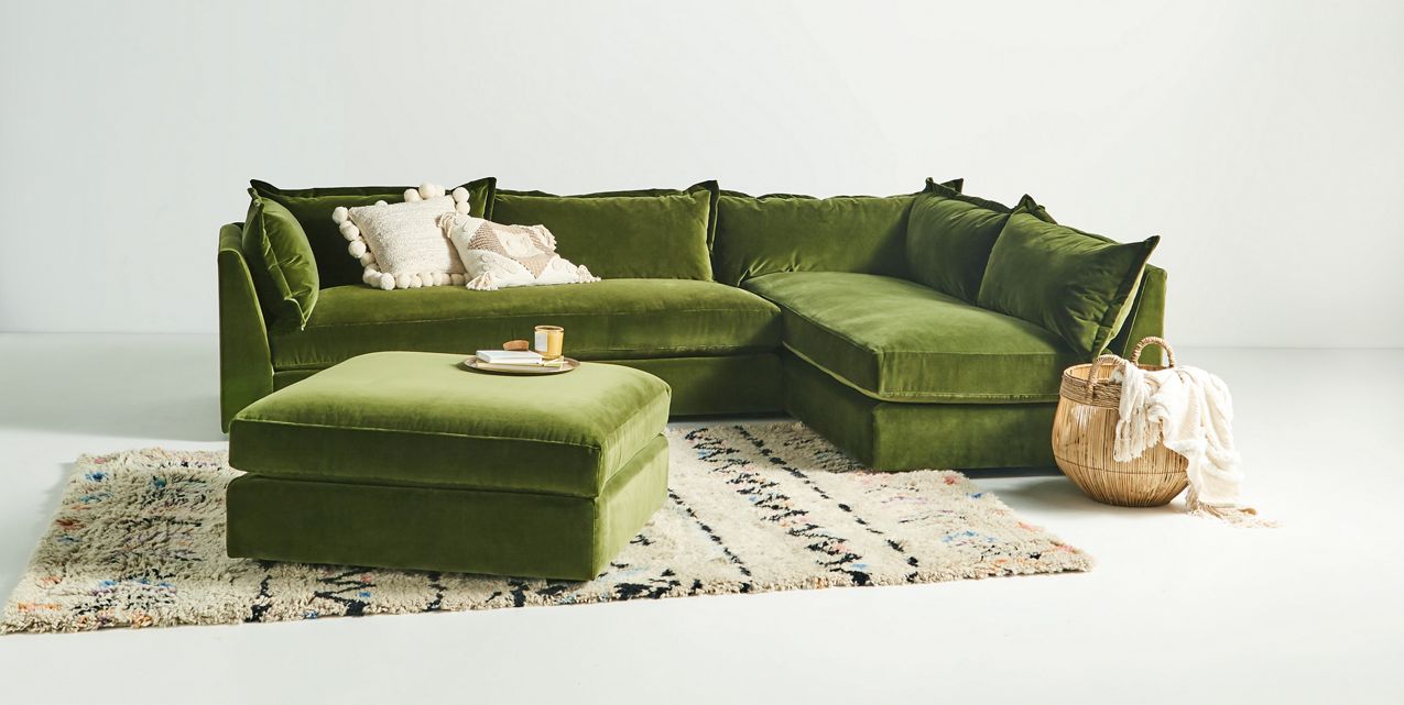 The Denver Two Piece Velvet L Shaped 119" Sectional in green is elegantly positioned on a patterned rug. A matching ottoman supports a decorative tray with candles, while a wicker basket housing a blanket sits nearby, crafting an inviting and cozy living area.