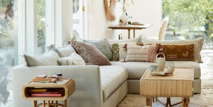 A cozy living room features the Denver Two Piece Velvet L Shaped 119" Sectional in light gray, embellished with a mix of patterned cushions. A small wooden side table is home to books and a candle, as sunlight pours through large windows that offer a view of the garden. A desk can be seen in the background.