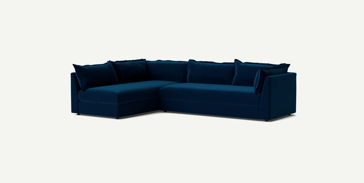 A Denver Two Piece Velvet L-Shaped 119" Sectional in dark blue with plush cushions is set against a plain off-white background. The sofa features a modern design with clean lines and a sleek appearance.