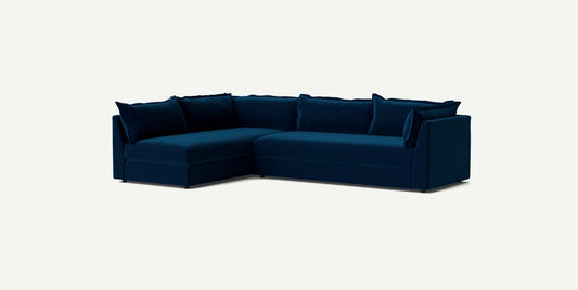 A Denver Two Piece Velvet L-Shaped 119" Sectional in dark blue with plush cushions is set against a plain off-white background. The sofa features a modern design with clean lines and a sleek appearance.