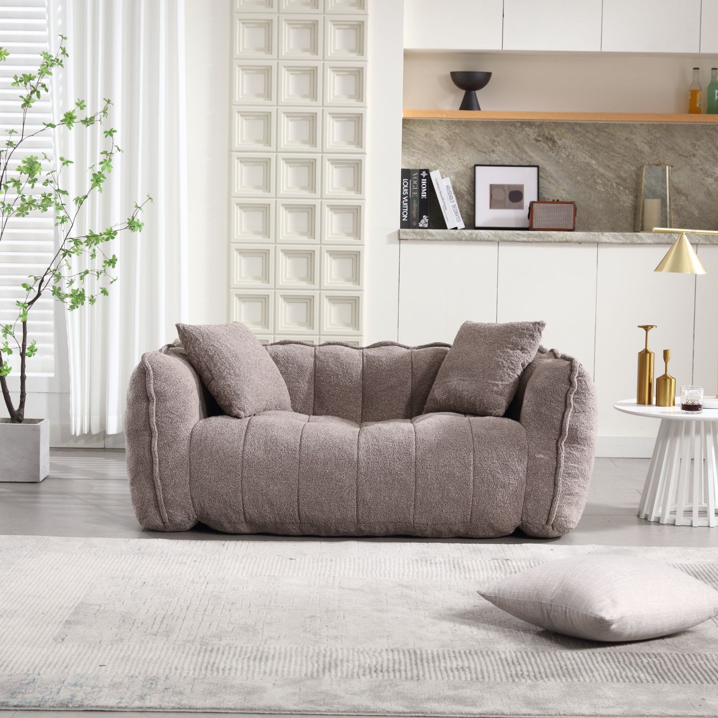 In a modern living room, the soft beanbag chair with a high resilience foam core for two people sits against textured walls. A marble shelf holds decor, and light floods through blinds on the left. A side table with books and a lamp is nearby atop a beige rug and pillow.