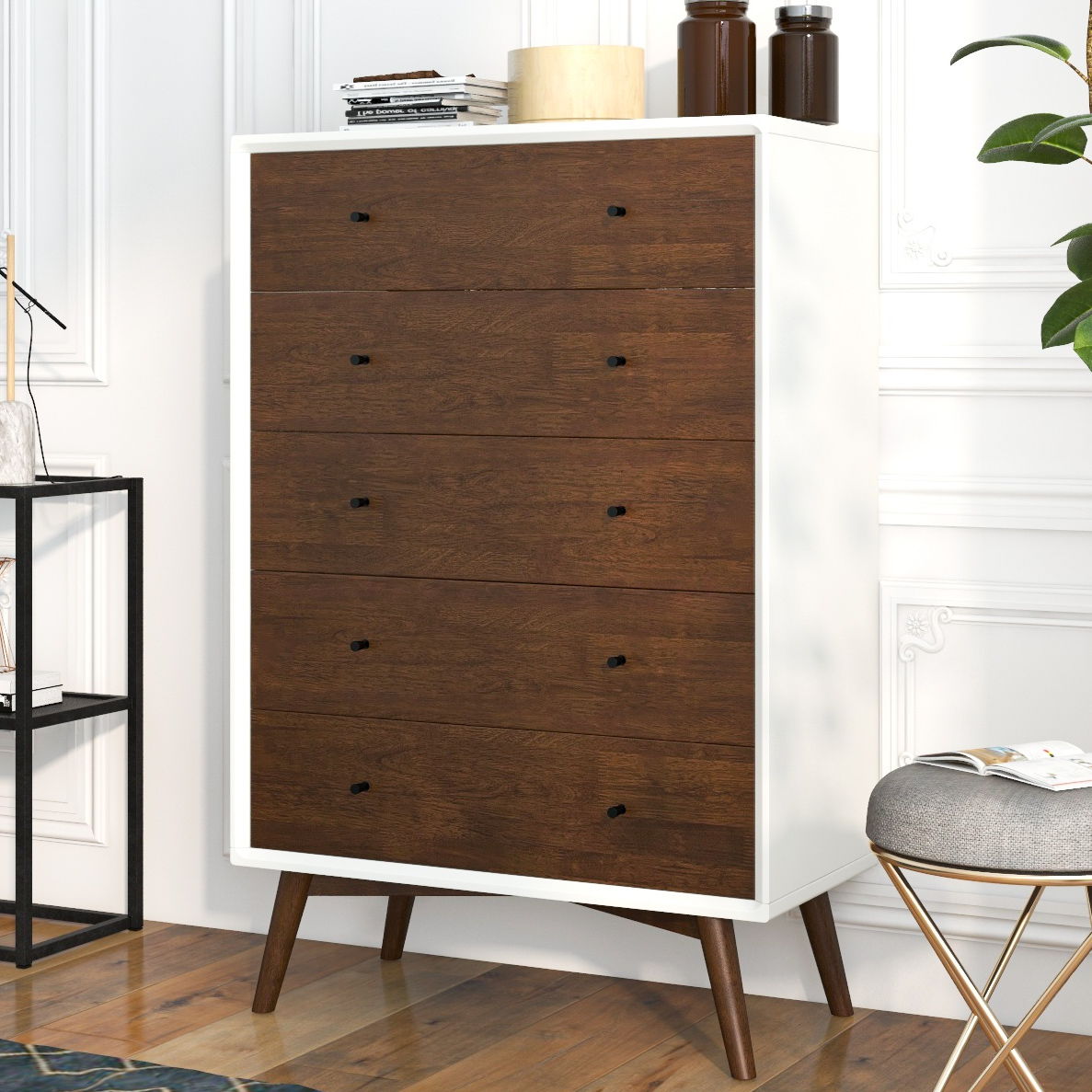 The Caroline dresser features a tall, mid-century modern design with a white frame and five wooden drawers accented by black knobs. It stands on angled legs and is topped with a lamp, books, and bottles. Nearby are a stool and black metal shelf on the wooden floor.