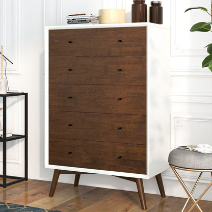 The Caroline dresser features a tall, mid-century modern design with a white frame and five wooden drawers accented by black knobs. It stands on angled legs and is topped with a lamp, books, and bottles. Nearby are a stool and black metal shelf on the wooden floor.