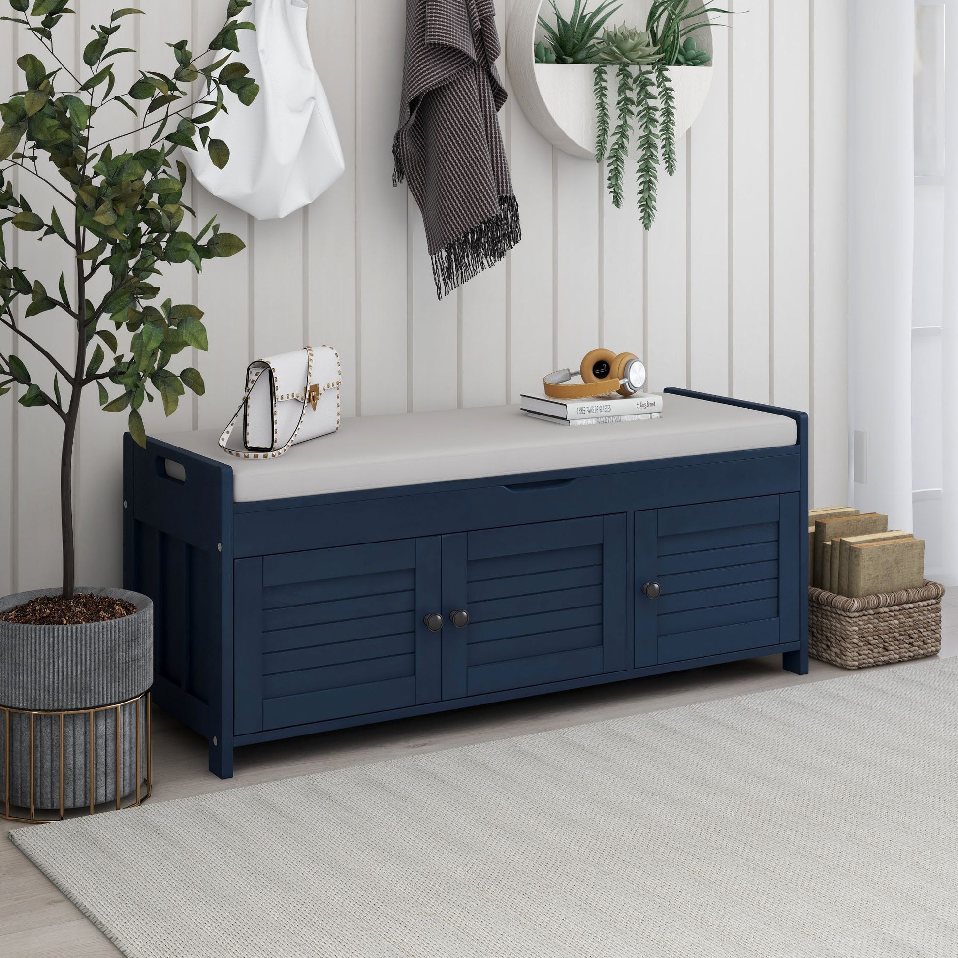 A navy blue storage bench with three shutter-shaped doors and a beige removable cushion features side handles and hidden storage. On top, it holds a white handbag, books, and a camera. Nearby decor includes a hanging mirror with plants, a scarf, coat, and basket with plant.