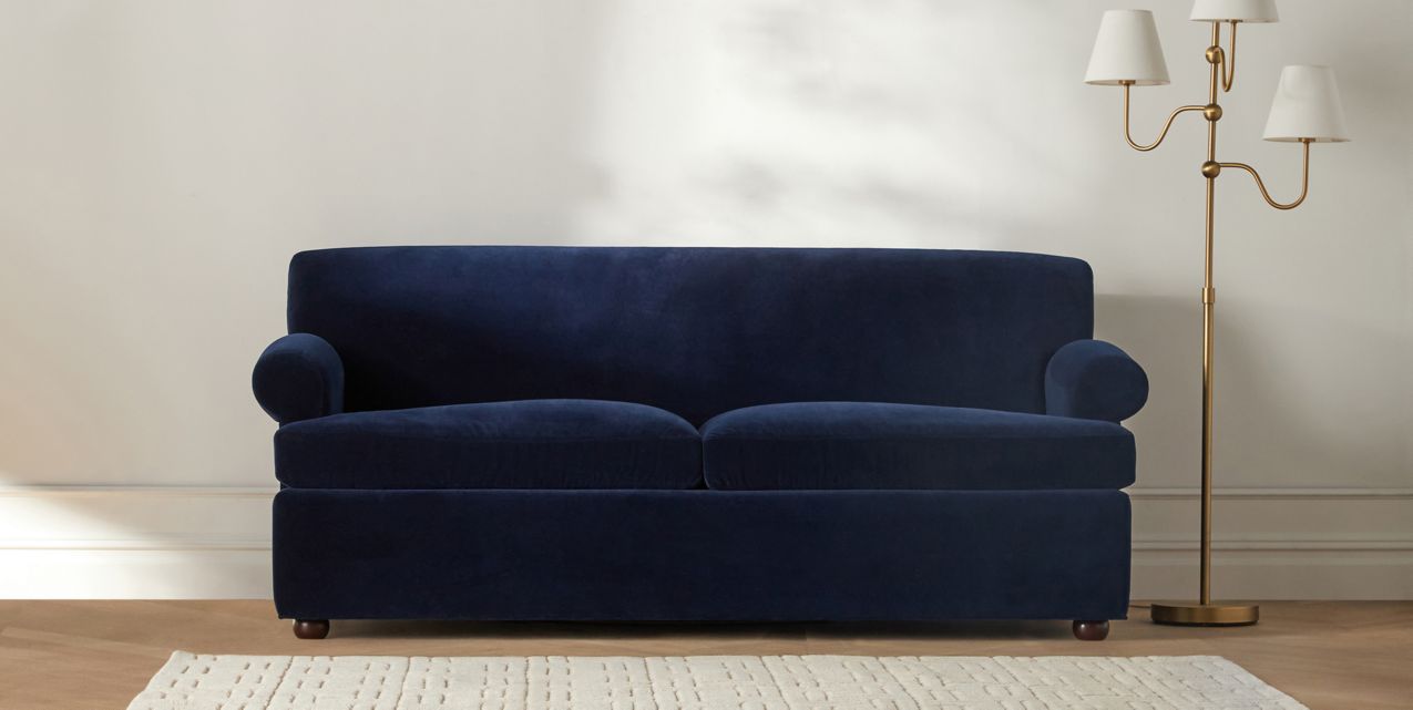 The Willoughby Sleeper Sofa in dark blue velvet, featuring rounded armrests, sits against a light-colored wall. A white textured rug is on the floor in front of the sofa. To the right, there is a tall gold floor lamp with three white lampshades.