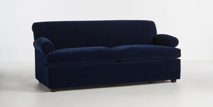 The Willoughby Sleeper Sofa, featuring plush cushions and rounded armrests in a rich dark blue hue, is set against a plain white background. Its sleek, modern aesthetic is accentuated by subtle wooden legs.