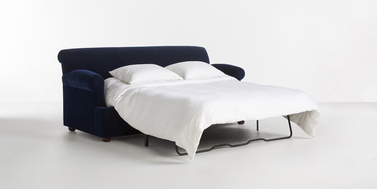 The Willoughby Sleeper Sofa in dark blue effortlessly converts into a double bed, complete with two white pillows and a white duvet. Set against a plain light gray backdrop, it showcases the modern and practical design of this versatile furniture piece.