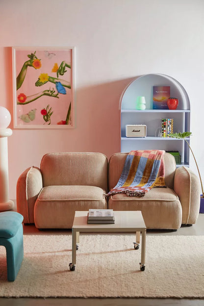 A cozy living room features the Macy Modular Sofa, draped with a colorful striped blanket. In front, a small table stands atop a beige rug. A blue arched shelf and vibrant abstract artwork adorn the wall, adding color to the space.