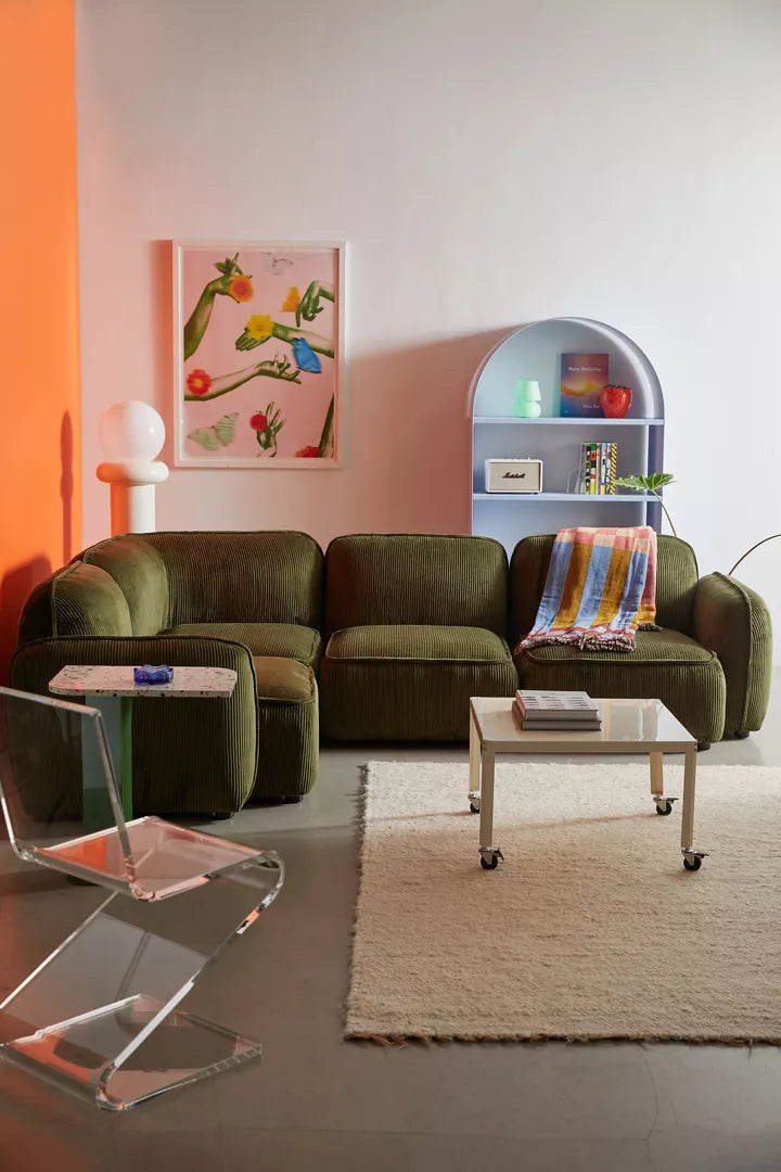 A modern living room featuring the Macy Modular Sofa in olive green, paired with a transparent acrylic chair and a white rug on the floor. A colorful painting is displayed on an orange wall. A white shelf showcases decorative items, while a small table draped with a scarf sits in front of the sofa.