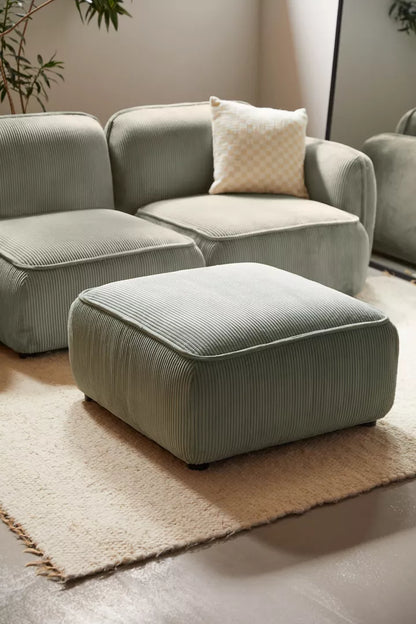 A cozy living room corner showcases the Macy Modular Sofa in green corduroy, accompanied by a matching ottoman on a light rug. A beige checkered pillow rests on the sofa, while soft lighting and a few plants enhance the serene atmosphere.