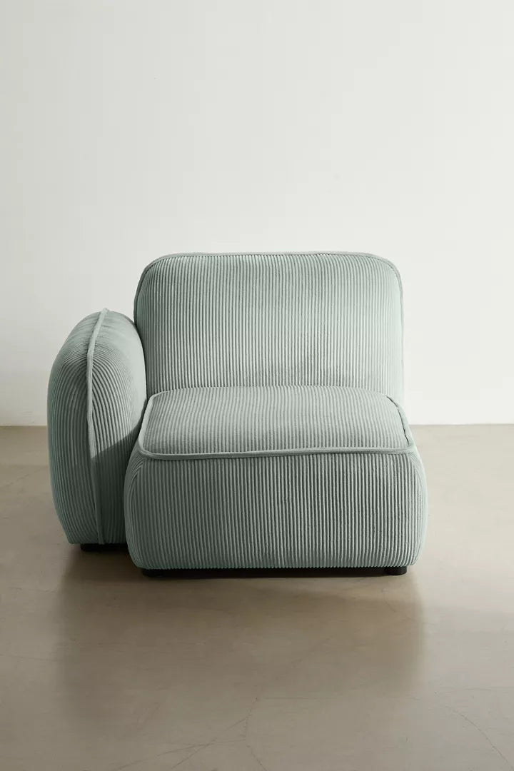 The Macy Modular Sofa features a modern design with armless sections, covered in a soft, ribbed fabric texture in a muted green hue. This piece rests on a polished concrete floor and stands against a plain white wall, evoking a minimalist and serene ambiance.