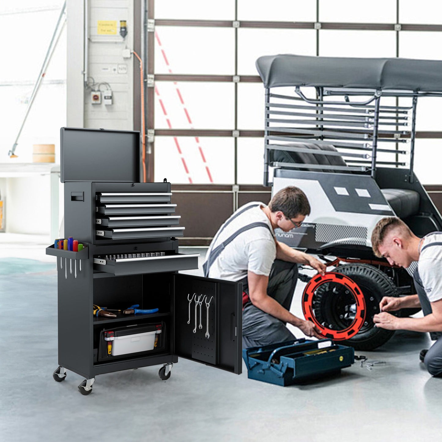 Two people work on a vehicle's tire inside a garage. A black 5-drawer rolling tool chest with open drawers is nearby, containing tools. One person kneels with tools, and the other holds the tire. A large garage door looms in the background.