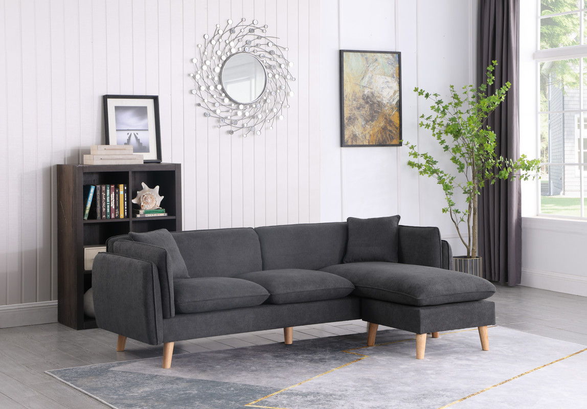 A modern living room features the Brayden fabric sectional sofa chaise in dark gray on a light gray rug. White paneled walls hold a circular mirror and artwork. A black bookshelf with books and decor sits left, while a potted plant stands right near a window with dark curtains.