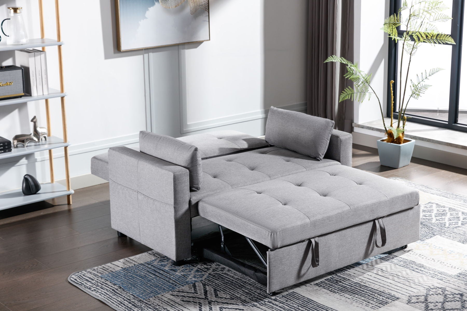 The 2-Seater Sleeper Sofa Bed in modern gray linen fabric is extended into a bed form in a bright room. It has two cushions, sits on a patterned rug, flanked by shelves with decor to the left, and is next to a tall window with a potted plant nearby.