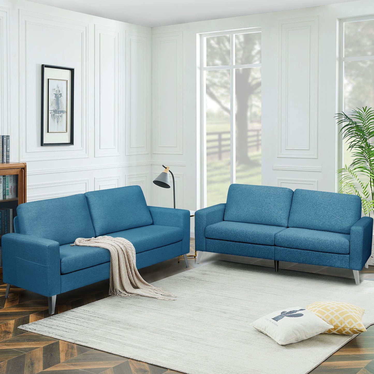 A living room featuring two YODOLLA 79" Sectional Sofas in blue, placed on a white rug, accompanied by a floor lamp, a potted plant, and a framed picture on the wall. A soft throw is elegantly draped over one of the sofas. Large windows reveal a view of trees outside.