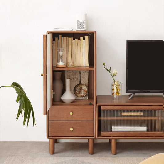 A Free-Standing Corner Cabinets Storage Table in walnut with vintage glass door features 2 shelves displaying books, an hourglass, and vase. Below are 2 drawers with brass handles. A TV sits on a matching stand with a plant and vase beside it, while a green leafy plant leans in from the left.