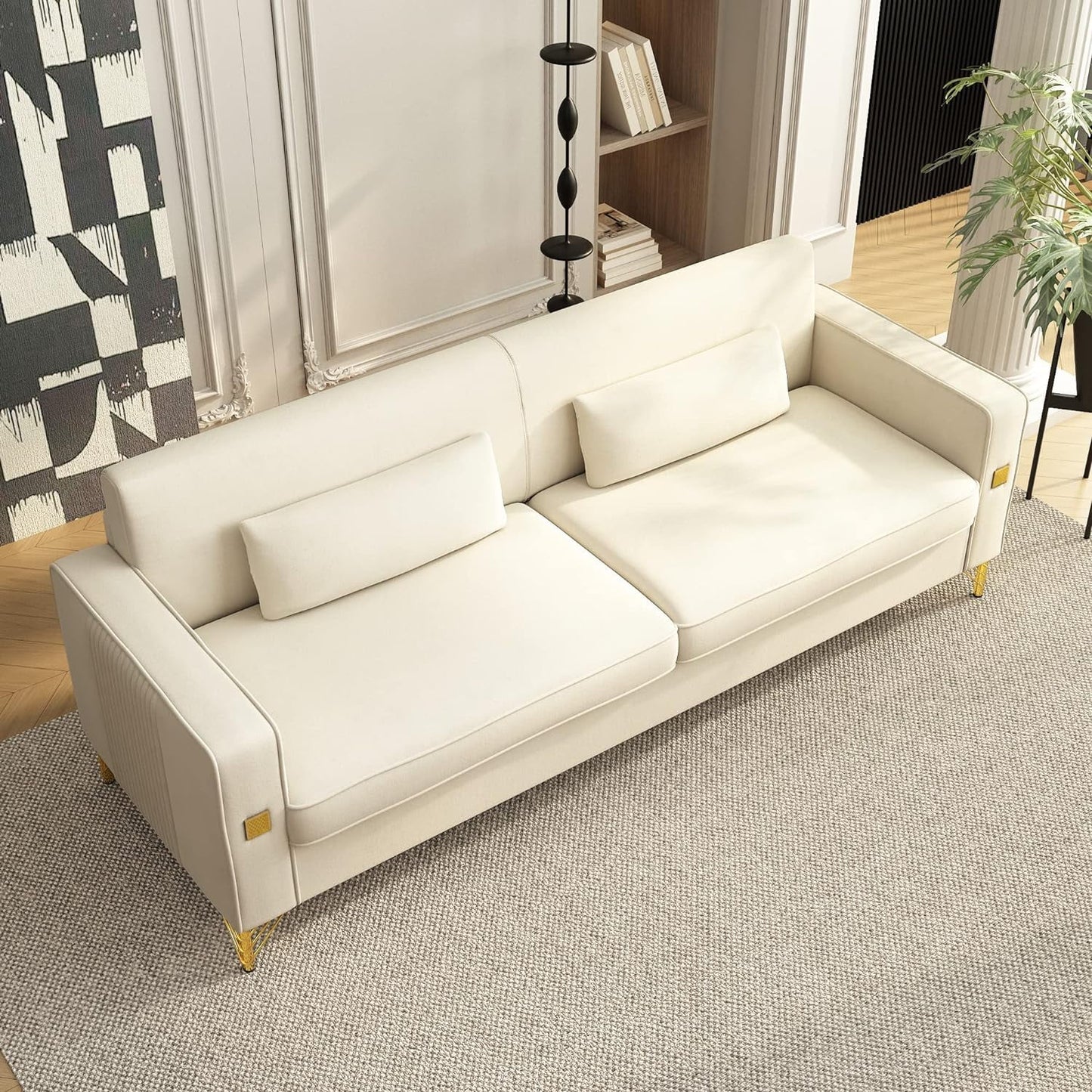 A sleek, modern Mid Century Modern Sofa Couch with two cushions is placed on a light gray textured rug in a stylish room. In the background, a tall black floor lamp and a bookshelf with a few books can be seen, complemented by a leafy plant visible on the right side.