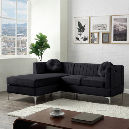 A modern living room features the Chloe Velvet Sectional Sofa Chaise with a USB charging port and two round cushions. Framed art adorns the wall, while a potted plant sits beside the sofa. A large window displays a cityscape view, and a light rug lies near a wooden table with books.