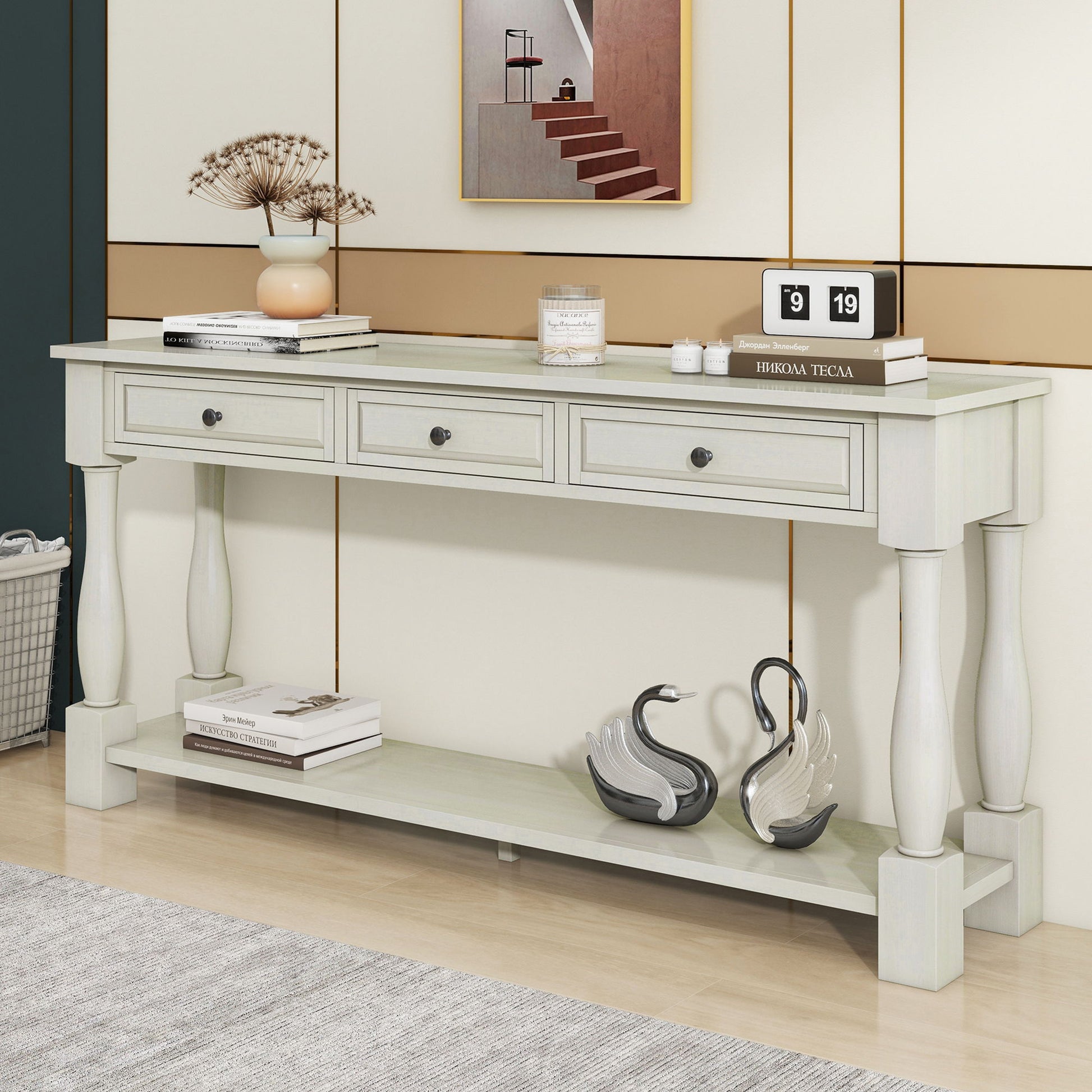 A Console Table Long Console Table with Drawers and Shelf displays books, a candle, and an ornate plant on the top. Two swan-shaped figurines are on the lower shelf. Behind it, there is a framed picture and a retro calendar on the wall.