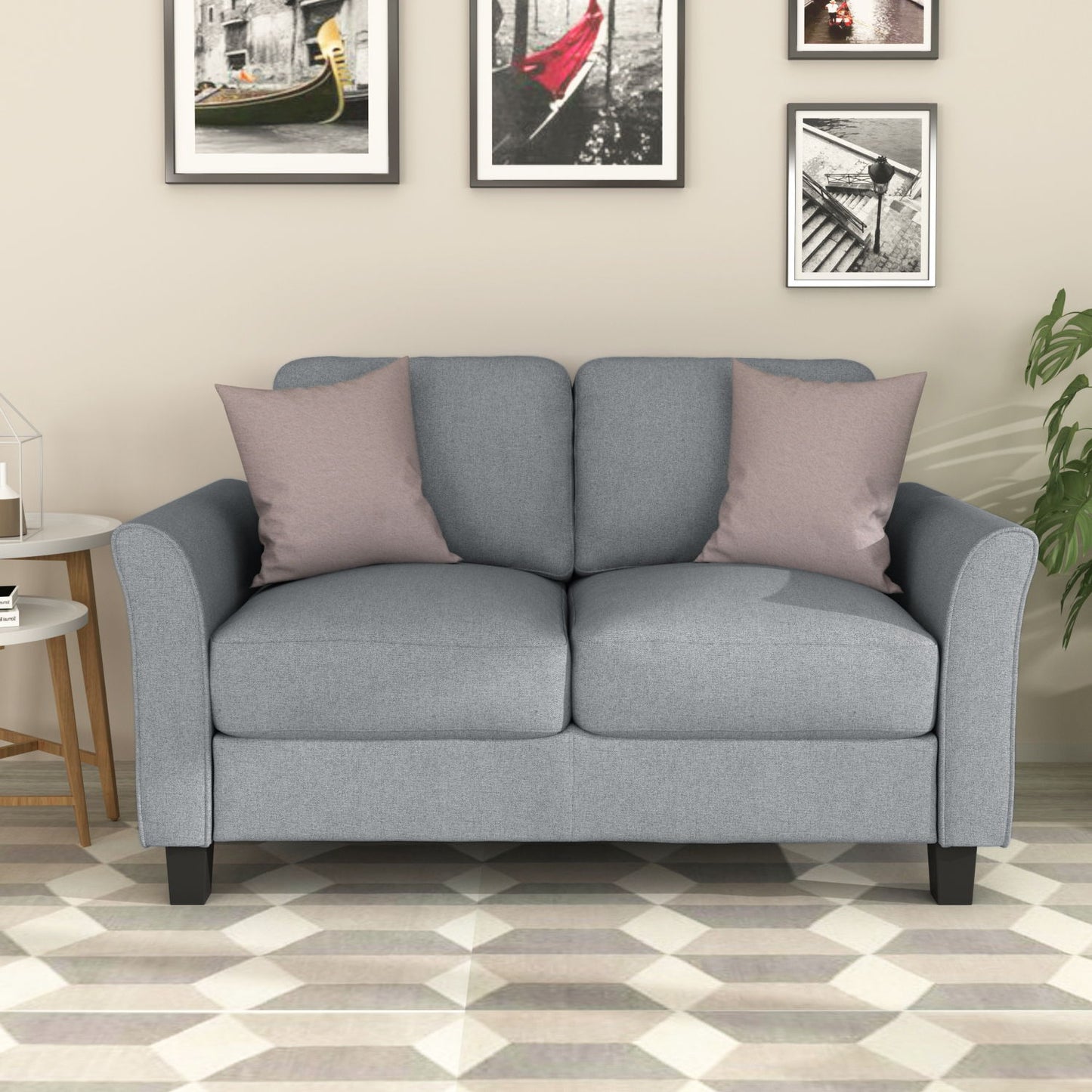 A light gray loveseat sofa, accessorized with two pink cushions, stands against a beige wall displaying black-framed photos. On the left is a white side table with decor items, while on the right sits a green potted plant. The floor features a geometric pattern.