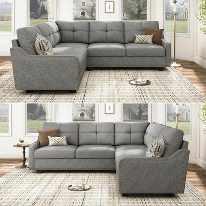 HONBAY Convertible Sectional Reversible Apartment sofa with tufted backrests and nailhead trim, set in a living room adorned with patterned throw pillows. It rests on a light-colored rug placed over a wooden floor. Large windows offer a view of trees and a stone building outside.