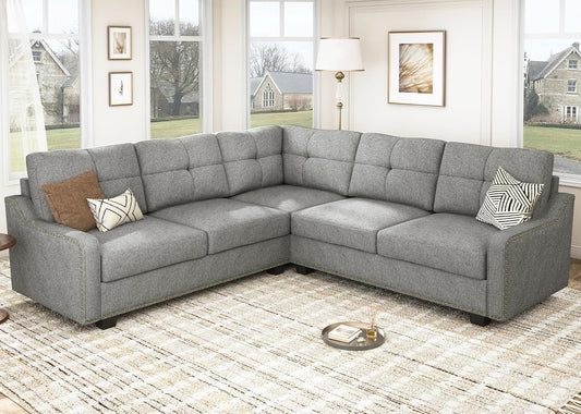 A cozy living room showcases the HONBAY Convertible Sectional Reversible Apartment sofa in gray, adorned with decorative pillows. The space includes a patterned rug, a floor lamp, and framed wall art. Large windows provide a view of the outdoors, revealing houses and trees in the distance.