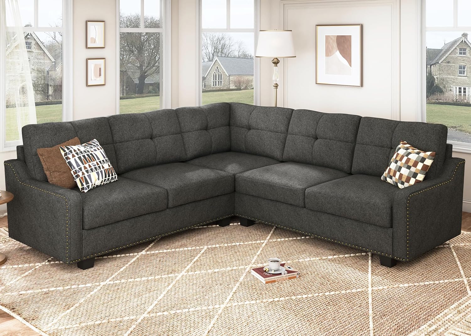 A modern living room features the HONBAY Convertible Sectional Reversible Apartment, a spacious gray sofa complemented by patterned cushions. Behind the sofa, a floor lamp adds illumination, while an abstract painting in a frame adorns the wall. The room boasts large windows and is grounded by a beige carpet with a geometric design.