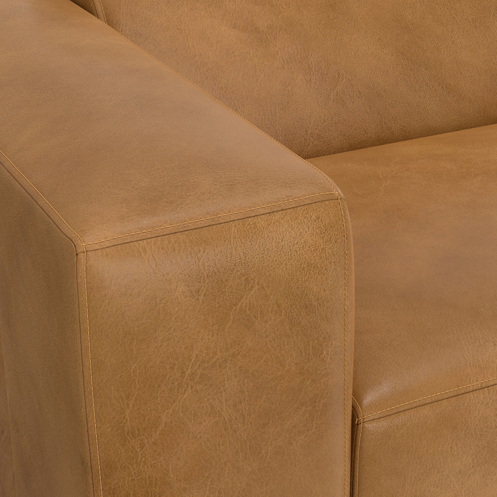 Close-up of a corner of a tan leather sofa with smooth stitching and a structured armrest. The texture of the leather is visible, showcasing its sleek and polished appearance.