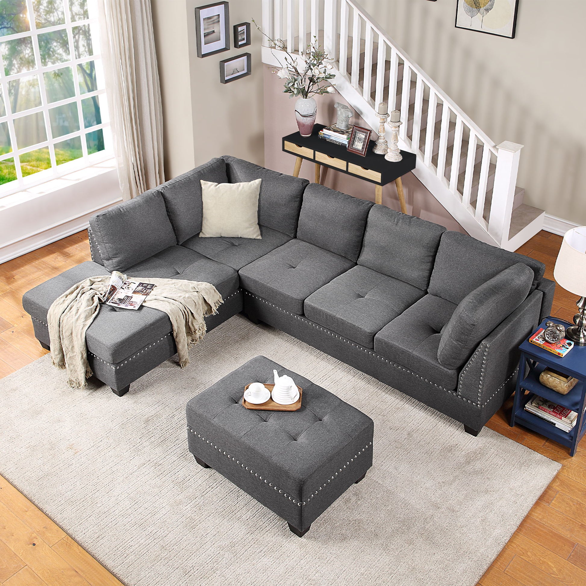 A cozy living room features a Reversible Sectional Sofa Space Saving L-Shaped Couch with a storage ottoman, all on a light beige rug. Sheer curtains allow natural light through the window, while a side table with a lamp and decorative staircase complete the space.