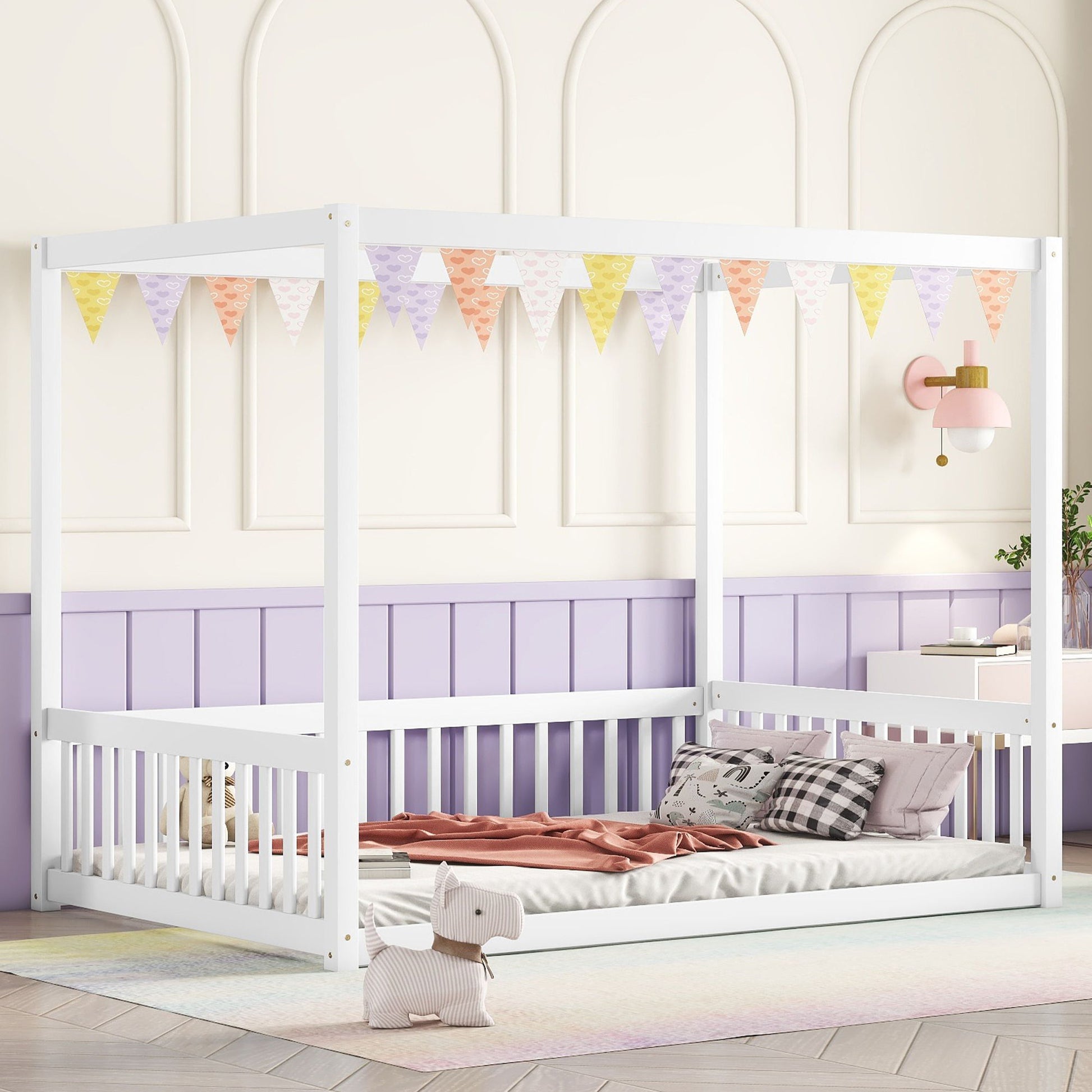 Against a half light purple wall, a white Canopy Frame Floor Bed with Guardrails for children is placed. Pastel bunting decorates above it, and a pink sconce adorns the wall. Nearby, on a soft multicolored rug, sits a plush toy horse.