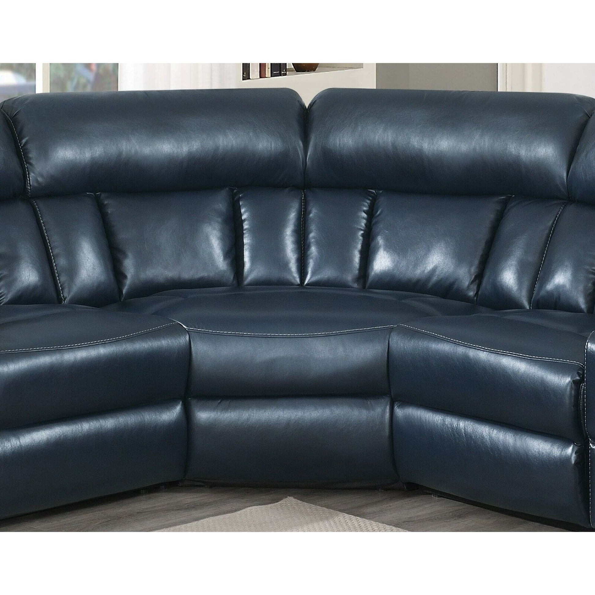 A Luxurious Manual Motion Sectional Sofa in dark blue leather, featuring plush cushions and a curved design, rests on a light-colored floor. The sofa's stitching details are noticeable, with a window and bookshelf partially visible in the background.