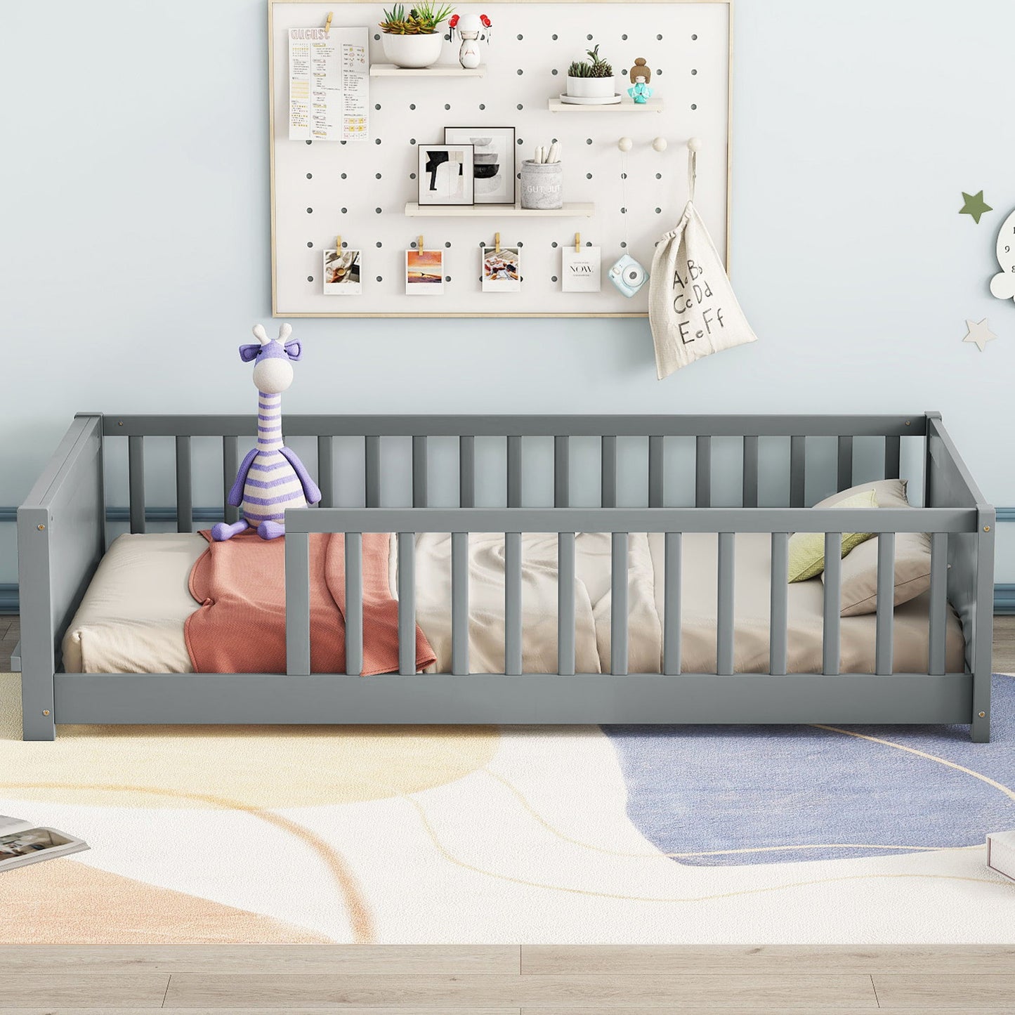 A child's room with a gray floor platform bed that includes built-in book storage. A striped purple giraffe plush toy sits on the bed. The wall features a pegboard with small plants, framed pictures, and a hanging string bag. A colorful abstract rug brightens up the floor.