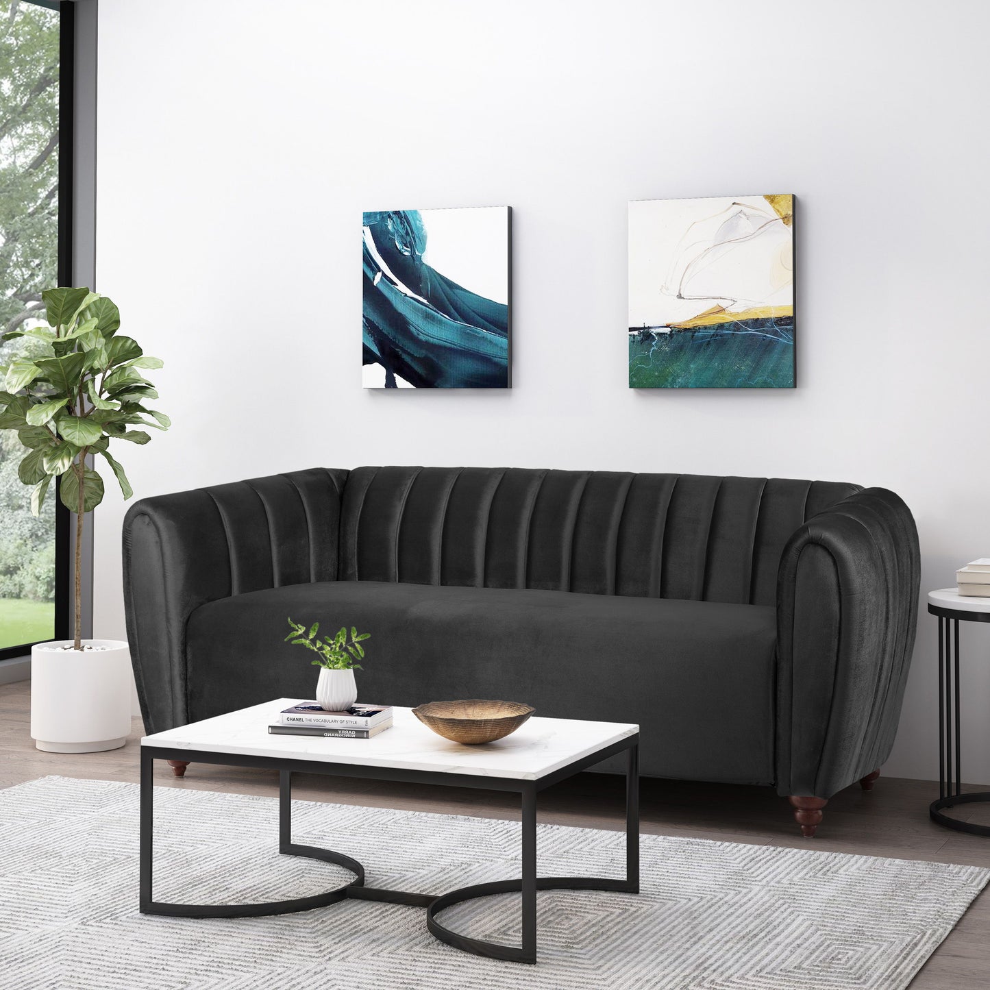 In a modern living room, a dark gray 3 Seater Sofa Modern Glam Design sits against a white wall, beneath two abstract paintings. A white coffee table with a black metal frame holds a plant and bowl. A leafy plant by the large window reveals verdant scenery.