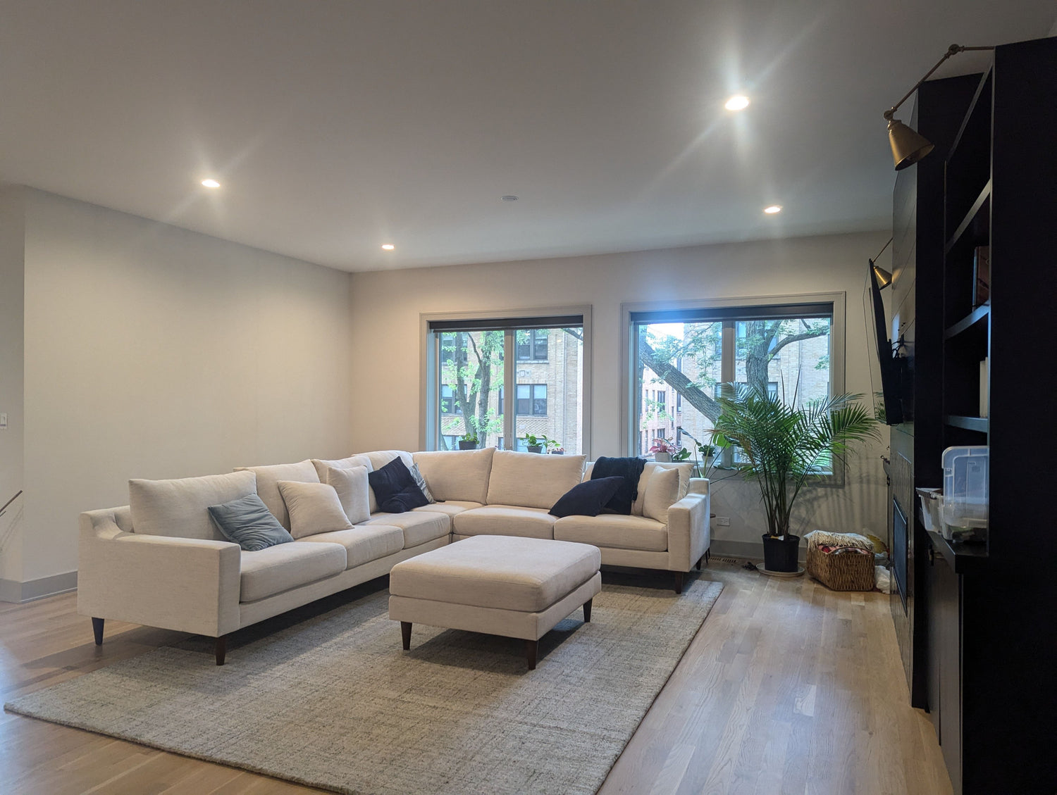 A modern living room with a large, light-colored sectional sofa and a matching ottoman on a textured rug. Large windows offer a view of trees outside. The room is decorated with plants and has a minimalist design.