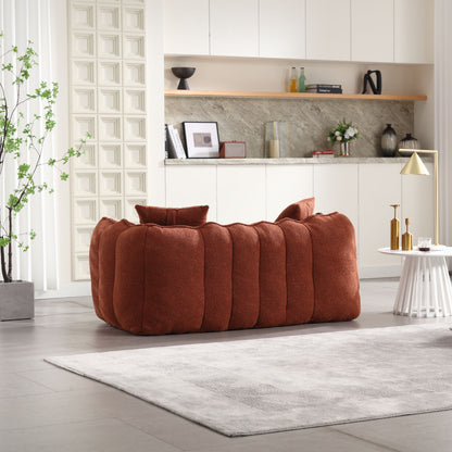 A contemporary living room featuring a plush, rust-colored beanbag chair for two with high-resilience foam. A round white side table, golden lamp, textured gray rug, minimalist kitchen, and tall plant provide stylish accents.