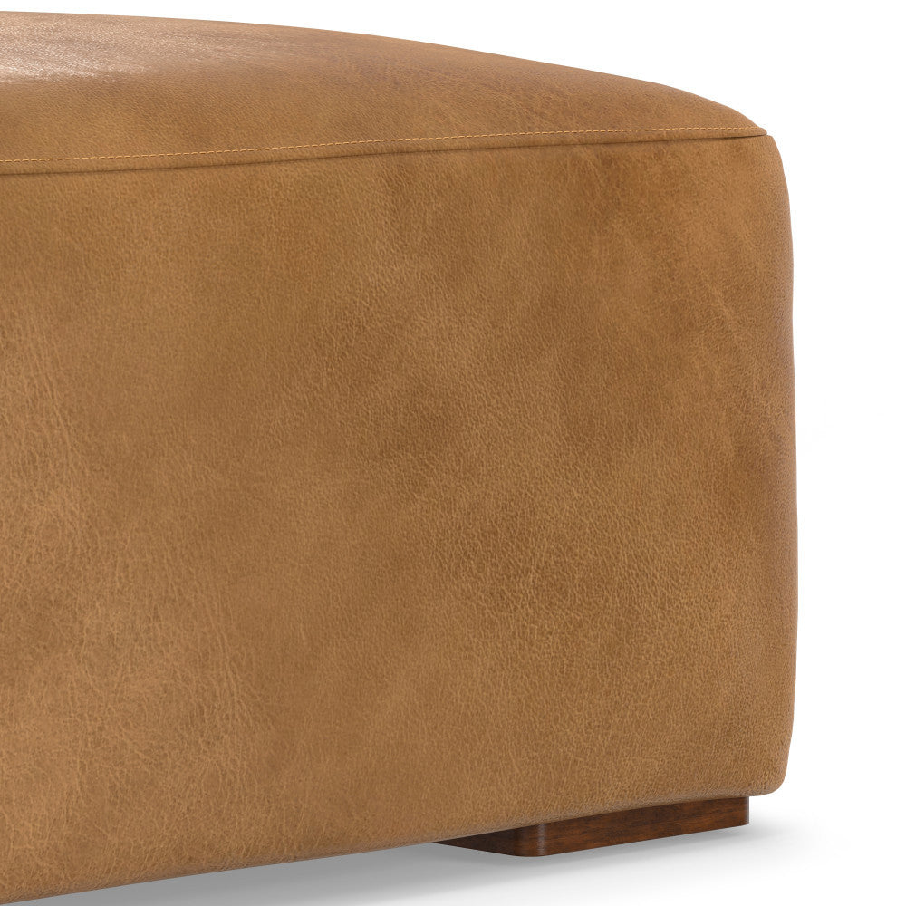 Close-up of a rectangular, light brown leather ottoman corner. The leather has a slightly textured surface. The ottoman sits on small, wooden block feet. The background is plain white.