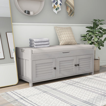 A Storage Bench with 3 Shutter-Shaped Doors featuring a removable cushion, hidden storage, and folded towels with a patterned pillow on top is beside a potted plant to the right; a geometric-patterned rug covers the floor.