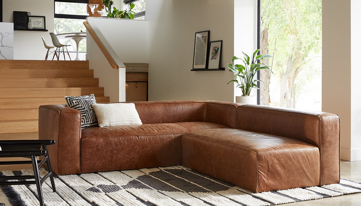 A modern living room with a brown leather sectional sofa on a patterned rug. Black and white throw pillows are on the sofa. Behind it, there's a staircase with wooden steps, potted plants, two framed artworks on the wall, and large windows letting in natural light.