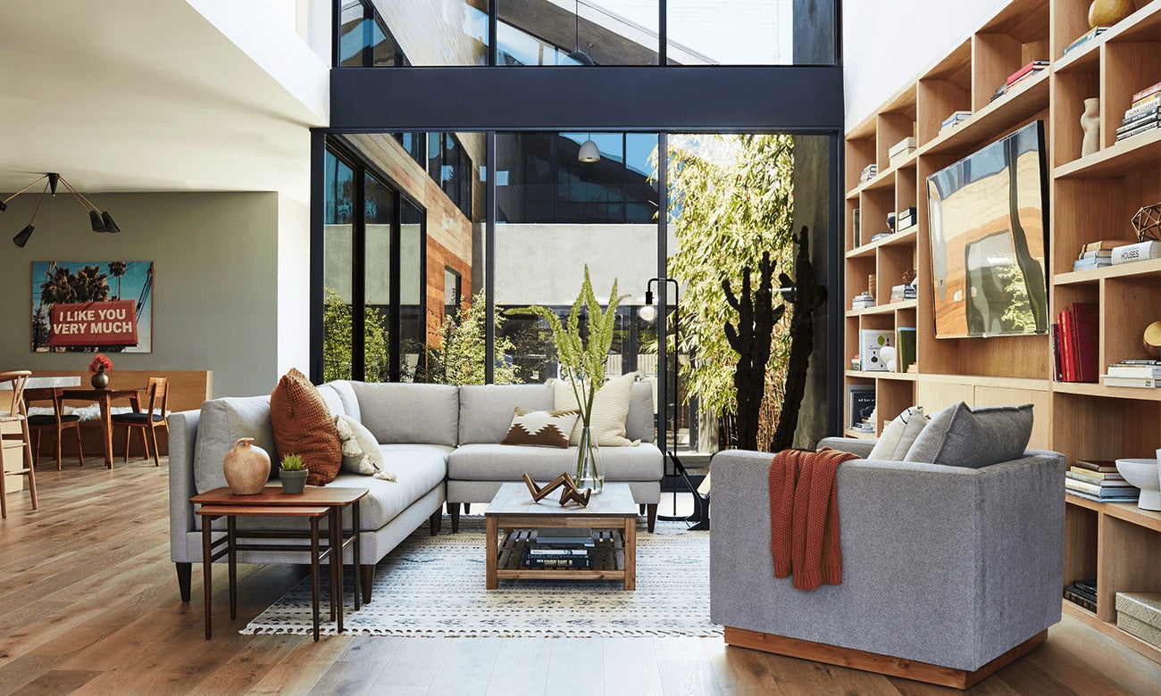 A modern living room with a large window showcasing an outdoor view. It features a gray sectional sofa, a gray armchair with an orange throw, a wooden coffee table, and open shelving filled with decorative items. In the background, there's a dining area with a wall art piece.