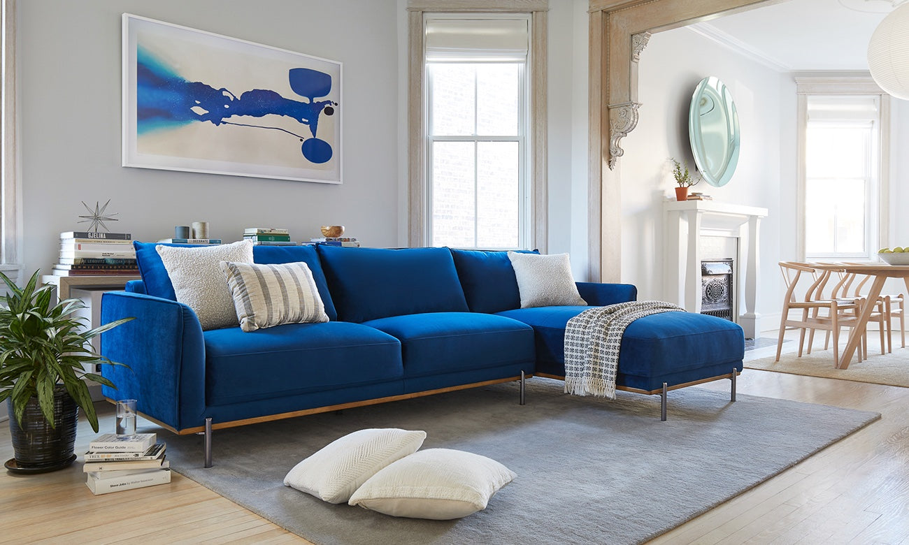 A modern living room with a blue sectional sofa adorned with white and gray pillows and a throw blanket. The room features a gray rug, light-colored wooden flooring, a plant, and abstract artwork on the wall. A dining area is visible in the background.