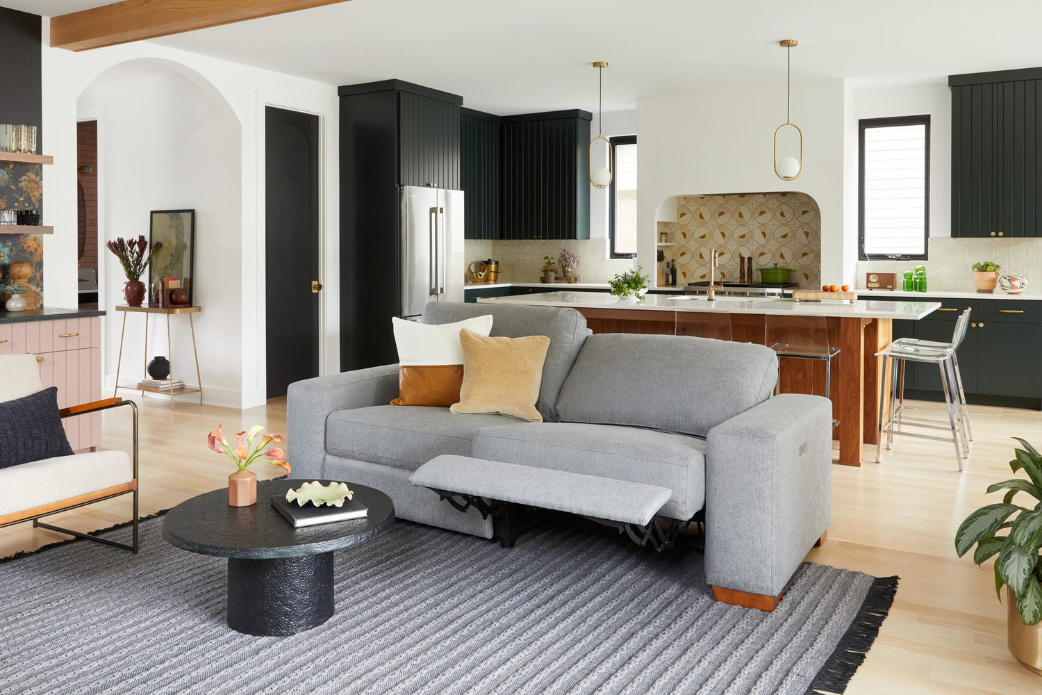 A modern living room and kitchen area with a gray reclining sofa adorned with pillows, a round black coffee table, light wood flooring, and black cabinets in the kitchen. Decorative items and plants enhance the cozy, contemporary ambiance.