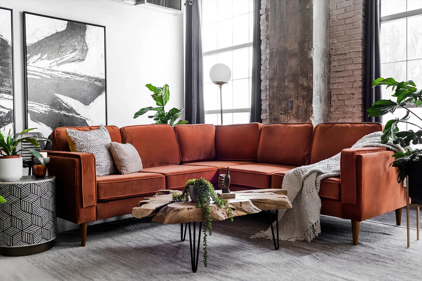 A modern living room featuring the Albany Corner Sectional in rust velvet, complemented by a wooden coffee table and various plants. Large windows and abstract artwork on the walls enhance the stylish decor, while a gray rug and brick walls complete the cozy space.