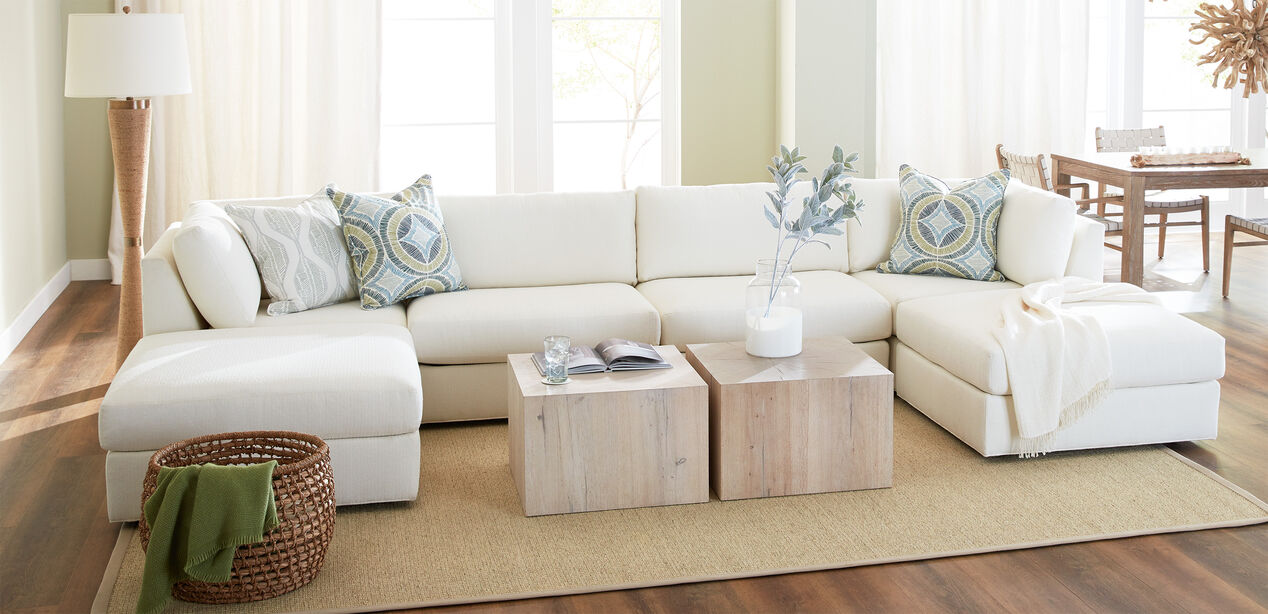 A cozy living room features the Daydreamer Modular Six-Piece Sectional, a large white sofa embellished with patterned throw pillows and a blanket. In front, two wooden coffee tables hold a book and a vase. On the floor, there's a woven basket along with a green throw, as sunlight streams through the windows.