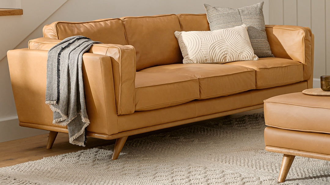 A modern tan leather sofa stands on a textured beige rug in a cozy living room. The sofa is adorned with patterned throw pillows in neutral tones and a gray knit blanket draped over the armrest. A matching ottoman is partially visible next to the sofa.