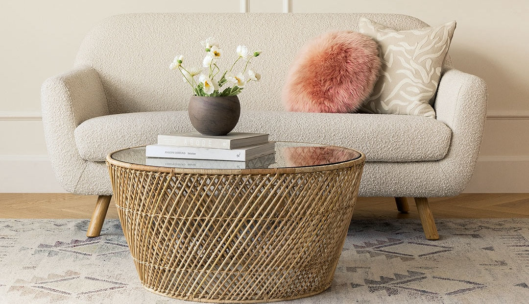 A cozy living room with a beige textured sofa, two decorative pillows (one pink fluffy and one with a beige pattern), and a rattan coffee table with a glass top. On the table are two books and a small vase with white flowers. A pastel patterned rug lies beneath.